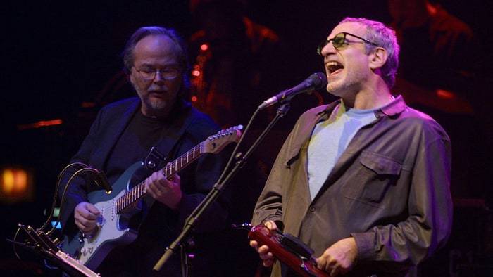 A man in sunglasses is singing into a microphone next to a man with a guitar.