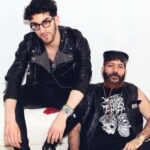 Two men in leather jackets sitting on a white couch.