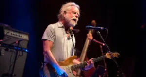 A man with a beard playing a guitar in front of a microphone.