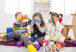 Three children sitting on a rug hold colorful plastic balls to their faces, surrounded by toys in a playroom.