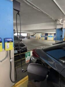 A car is parked in a parking garage and plugged into an electric vehicle charging station. Another car is parked in the adjacent space.