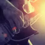 Close-up of a person playing an electric guitar with strong sunlight creating a lens flare in the background.