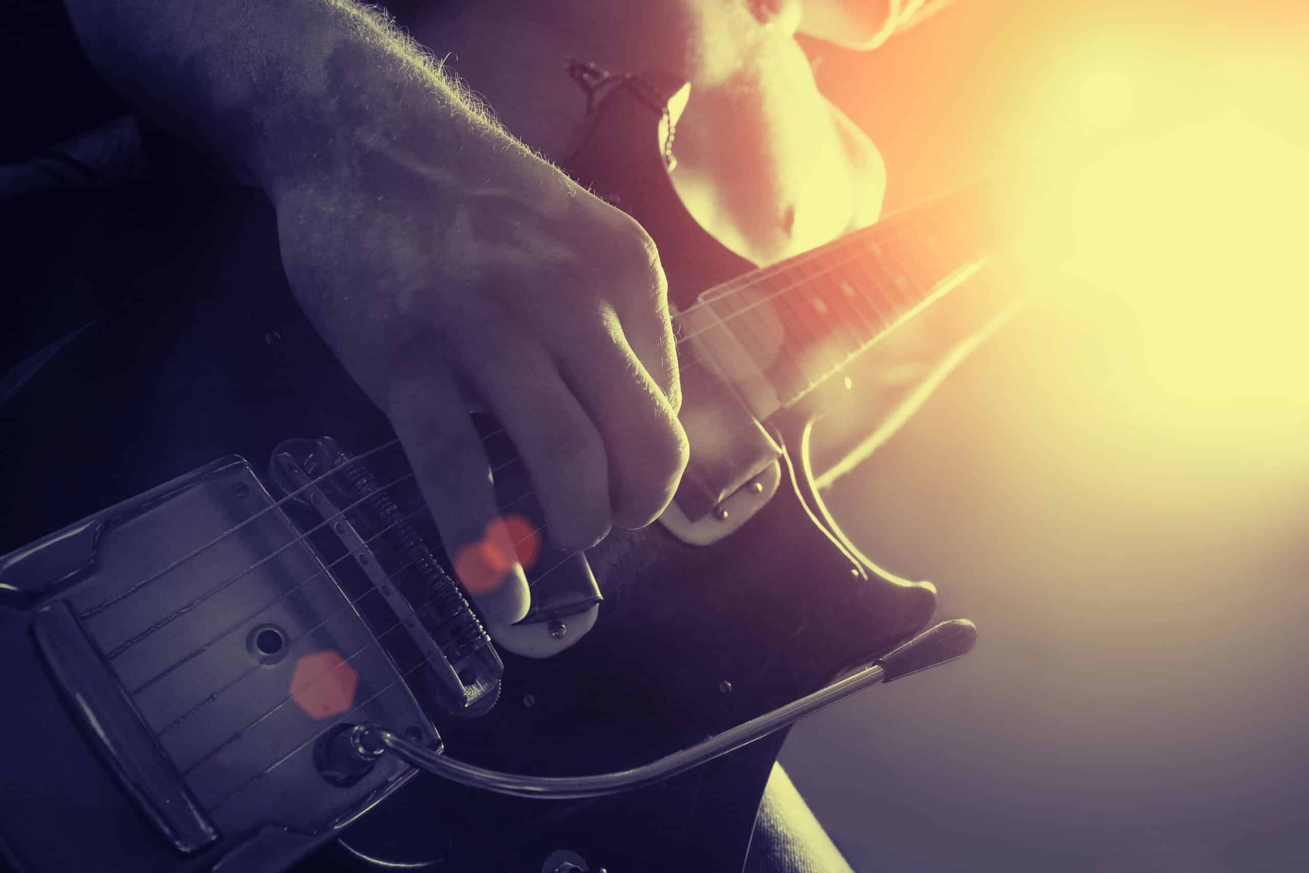 Close-up of a person playing an electric guitar with strong sunlight creating a lens flare in the background.