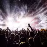 A crowd of people at a concert holds up phones and hands as bright stage lights and confetti fill the air.