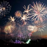 A large display of colorful fireworks explodes in the night sky above a dark landscape, with bright bursts and trails lighting up the scene.