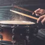 Close-up of a person striking a snare drum with drumsticks, causing particles to rise from the drumhead in dramatic lighting.