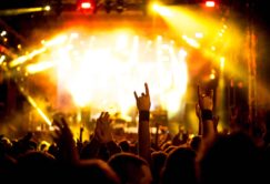 Crowd at a concert raising hands and making rock-and-roll gestures, illuminated by bright yellow stage lights.