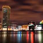 Night view of a city waterfront with tall buildings and a glass pyramid structure, colorful lights reflected on calm water under a cloudy sky.