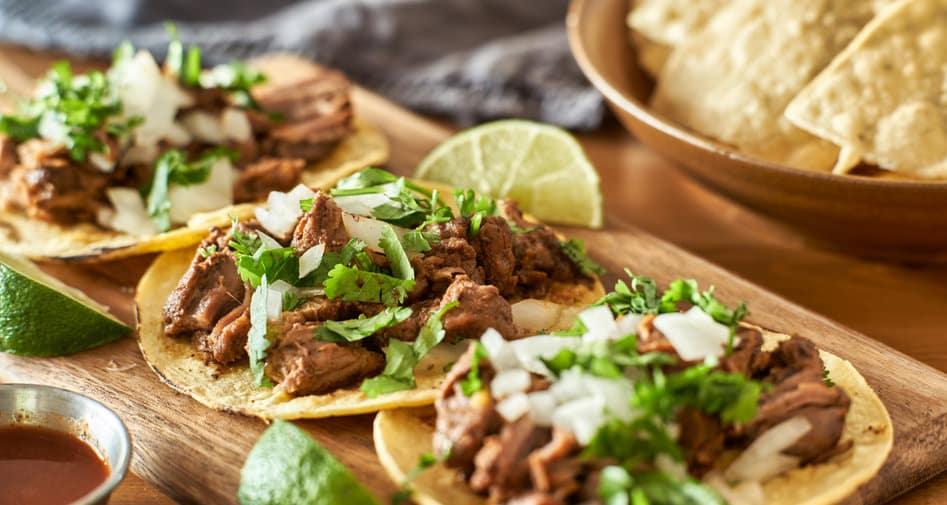 Three tacos topped with shredded meat, chopped onions, and cilantro on a wooden tray, with lime wedges and a bowl of tortilla chips in the background.