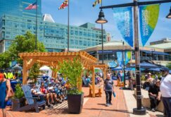 People sit and walk around a shaded seating area in an urban plaza with tall buildings, banners, and flags in the background on a sunny day.