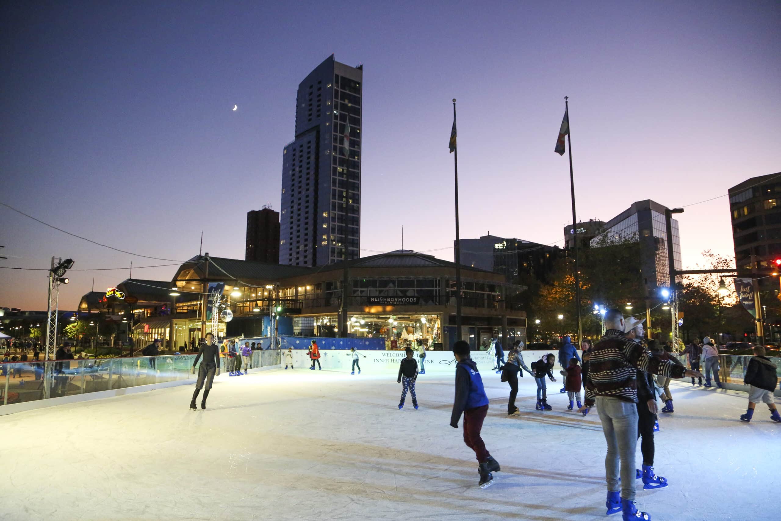 People ice skating at an outdoor rink in a city at dusk, with tall buildings and flags visible in the background under a twilight sky.