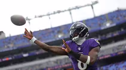 A football player in a purple Baltimore Ravens uniform throws a football on a stadium field with empty blue seats in the background.
