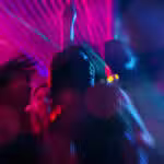 A group of people dance under colorful neon lights at a party, with blurred motion and vibrant pink and blue hues creating a dynamic atmosphere.