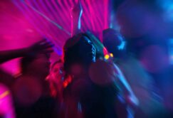 A group of people dance under colorful neon lights at a party, with blurred motion and vibrant pink and blue hues creating a dynamic atmosphere.