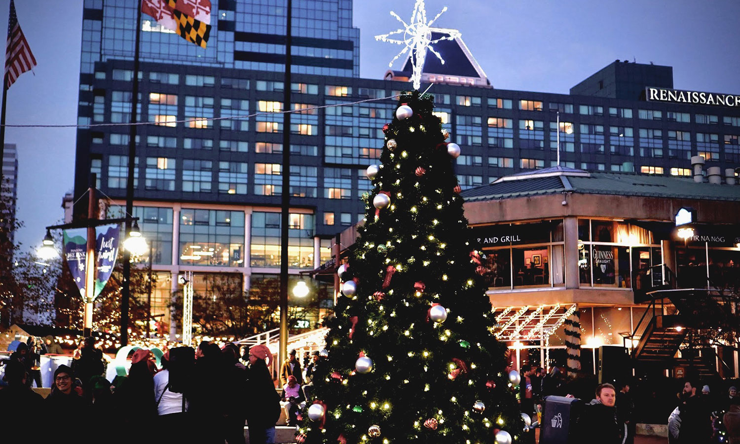 A decorated Christmas tree stands in a busy urban plaza at dusk, with people gathered around and city buildings in the background.