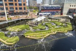 Floating wetlands with green plants are anchored in a harbor next to brick and modern buildings under a partly cloudy sky.