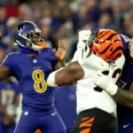 A football quarterback in a blue uniform prepares to throw the ball as a teammate blocks a defender in a white and orange uniform during a game.