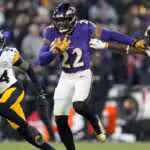 A football player in a purple Ravens uniform runs with the ball while fending off two defenders in white Steelers uniforms during a game.