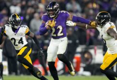 A football player in a purple Ravens uniform runs with the ball while fending off two defenders in white Steelers uniforms during a game.