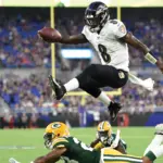 A Baltimore Ravens player leaps over a Green Bay Packers defender during a football game, holding the ball mid-air as other players watch.