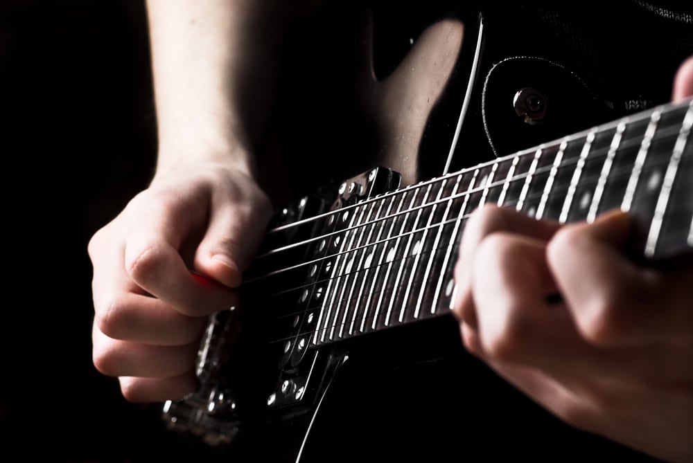 Close-up of a person playing an electric guitar, with one hand pressing the strings on the fretboard and the other hand strumming.