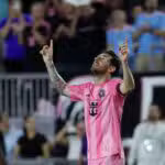 A soccer player, Messi, in a pink and black uniform raises both hands and points upwards during a match, with spectators visible in the background.