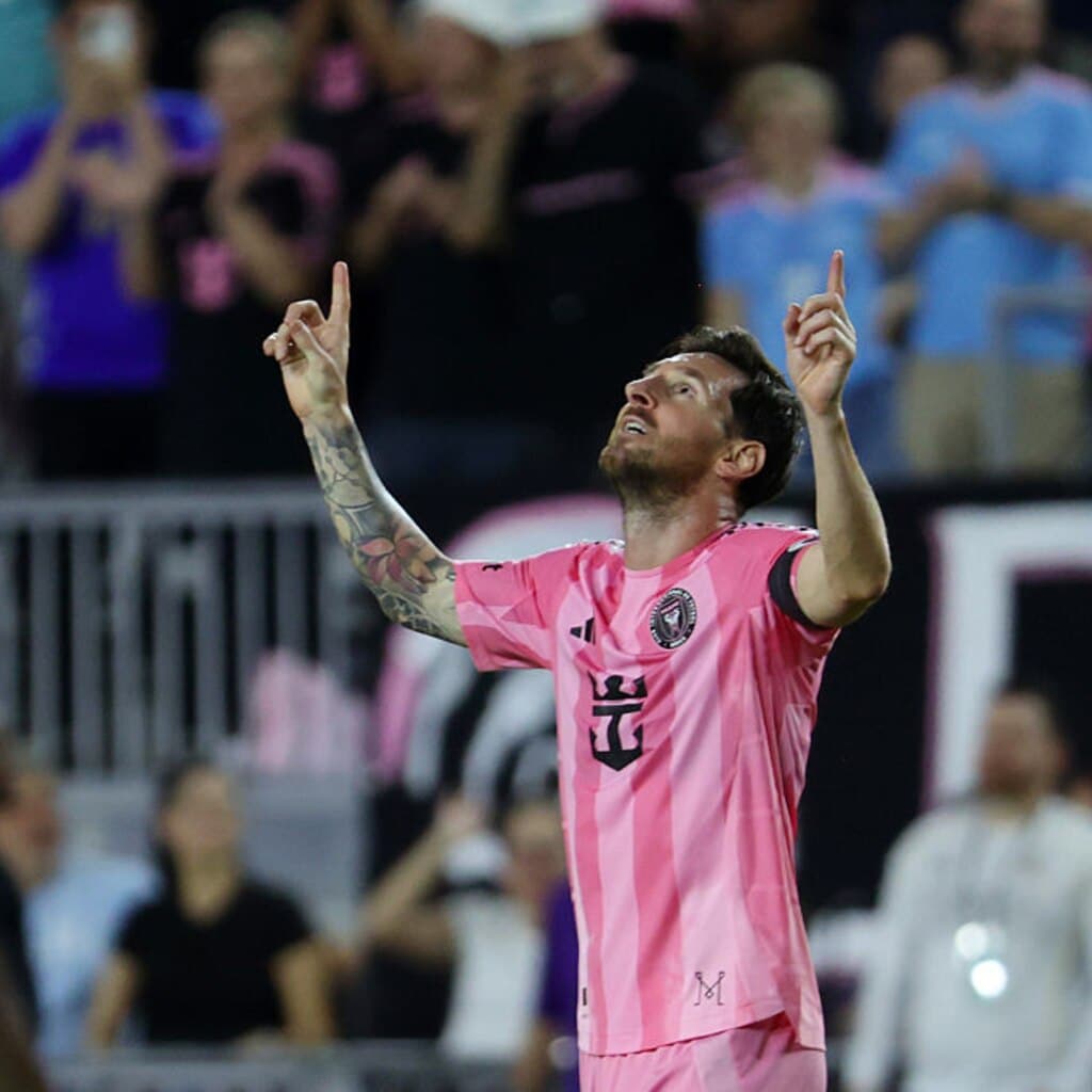 A soccer player, Messi, in a pink and black uniform raises both hands and points upwards during a match, with spectators visible in the background.